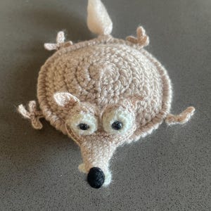 Könnte beinhalten: Eine beige gehäkelte Tierfigur mit einem kreisförmigen Körper, vier kleinen Gliedmaßen, einem spitzen Schwanz und einem Kopf mit zwei großen weißen Augen mit schwarzen Pupillen und einer schwarzen Nase. Die Garnstruktur ist im gesamten Design sichtbar.
