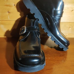 Può includere: Un paio di stivaletti in pelle nera con finitura lucida, punta quadrata e cinturino laterale fissato con una grande fibbia in metallo color argento. Gli stivali presentano una spessa suola in gomma nera con battistrada profondo per aderenza.