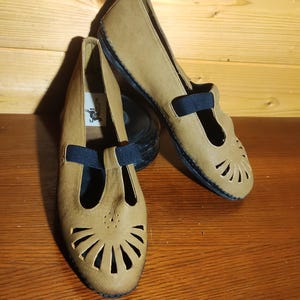 Può includere: Un paio di scarpe slip-on color marrone chiaro con un cinturino elastico nero sul collo del piede e un motivo decorativo traforato a ventaglio sulla punta arrotondata. Le scarpe hanno una suola nera e una piccola etichetta bianca all'interno.