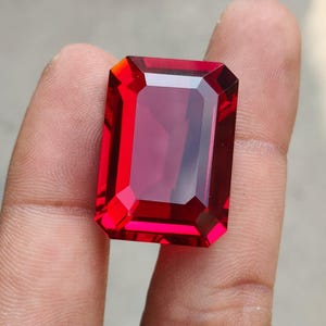 May include: A vibrant, deep red gemstone cut into a rectangular emerald shape with faceted edges. The stone exhibits high clarity and a polished surface, reflecting light across its transparent, saturated crimson body with sharp, defined corners.