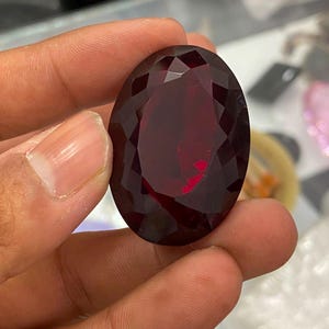May include: A large, oval-shaped, deep red gemstone with a faceted surface. The stone exhibits a dark crimson hue with light reflecting off its multiple polished facets, creating a brilliant appearance. The gem is held up to display its clarity.
