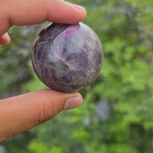 Può includere: Una pietra sferica lucida caratterizzata da un colore di base viola intenso con distinte e vivaci inclusioni magenta e rosa su tutta la superficie. La sfera presenta una consistenza liscia e riflettente con visibili fratture interne naturali.