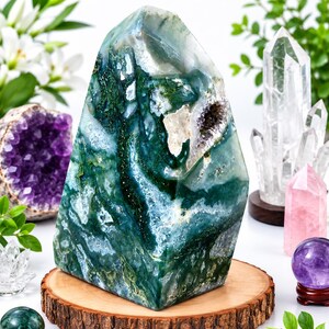 May include: A large, polished moss agate tower featuring deep green, translucent white, and grey mineral inclusions with a small, textured crystalline cavity on its side, displayed upright on a circular, natural wood slice base.