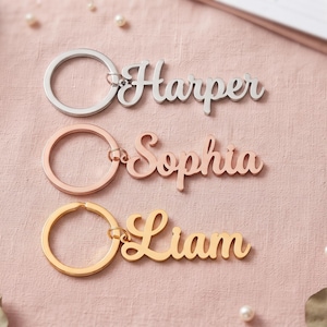 May include: Three metal keychains featuring custom names in a cursive script font. The top keychain is silver with the name Harper, the middle is rose gold with the name Sophia, and the bottom is gold with the name Liam, each attached to a ring.