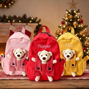 Könnte beinhalten: Drei Rucksäcke in Pink, Rot und Gelb, jeweils mit einem Plüschteddy in einer Vordertasche. Der pinke Rucksack ist mit Charlotte, der rote mit Harper und der gelbe mit Mia bestickt. Jeder Rucksack hat einen Tragegriff und schwarze Reißverschlüsse.