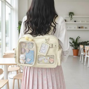 Könnte beinhalten: Ein cremefarbener Ita-Bag-Rucksack mit zwei durchsichtigen Fronttaschen, die mit verschiedenen kleinen Anstecknadeln, einer Plüschfigur und dekorativen Karten gefüllt sind. Der Rucksack verfügt über einen hängenden Ausweishalter und Seitentaschen.
