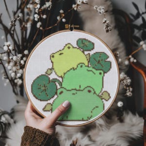 May include: A circular wooden embroidery hoop containing a cross-stitch pattern on white fabric. The design features three stacked green frogs in varying shades of lime and forest green, surrounded by several dark green lily pads.