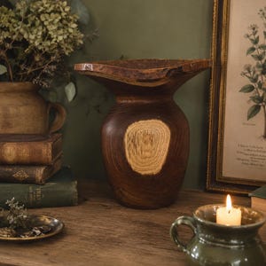 May include: A dark brown wooden vase with a wide, flared rim and a natural live edge. The body of the vase features a prominent, lighter-coloured circular wood grain pattern on the front, showcasing the natural texture and rings of the wood.