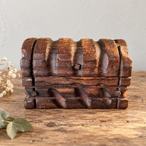 May include: A small, rectangular wooden chest with a dark brown finish and a curved, arched lid. The exterior features carved vertical grooves and horizontal decorative bands, with small square wooden pegs visible on the front surface.