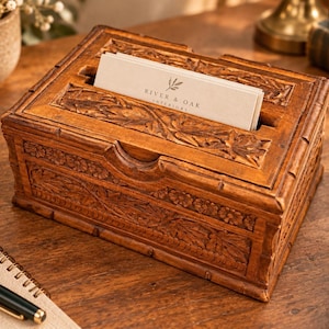 Puede incluir: Una caja rectangular de madera con intrincados tallados florales en los laterales y la tapa. La caja presenta una ranura central que contiene una pila de tarjetas de visita blancas impresas con el texto River & Oak Interiors y un pequeño icono de hoja.