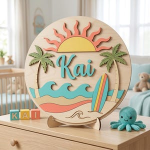 May include: A circular wooden sign featuring a layered design with a yellow sun, green palm trees, teal waves, a tan sandy beach, and a colourful surfboard. The name Kai is written in large teal script in the centre. The sign rests on a wooden stand.