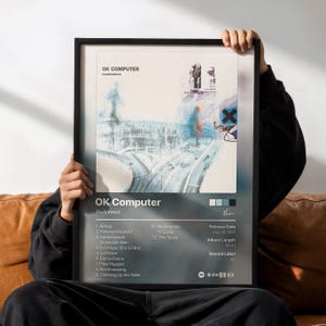Peut inclure: Une affiche encadrée présentant l'album OK Computer de Radiohead. L'illustration montre une scène d'autoroute aux tons bleus avec un symbole X noir. La liste des titres, de Airbag à The Tourist, figure en dessous avec les détails.