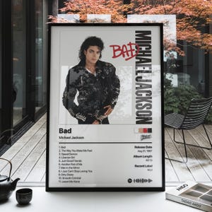 Puede incluir: Un póster enmarcado con la portada del álbum Bad de Michael Jackson. El diseño incluye un retrato del artista, el título BAD en rojo y una lista de canciones como Man in the Mirror y Smooth Criminal. La fecha de lanzamiento es el 31 de agosto de 1987.