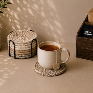 Pode incluir: Um conjunto de bases para copos redondas de corda trançada em tons neutros de bege e castanho, empilhadas num suporte de metal preto. Uma caneca de cerâmica salpicada com chá repousa sobre uma base, com uma etiqueta de chá English Breakfast.