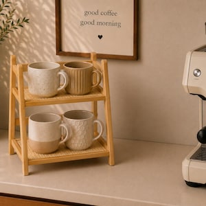 Può includere: Un espositore a due livelli in legno chiaro con base intrecciata, contenente quattro tazze in ceramica. Il ripiano superiore presenta una tazza bianca maculata e una tazza beige a coste. Il ripiano inferiore contiene una tazza bicolore e una bianca.