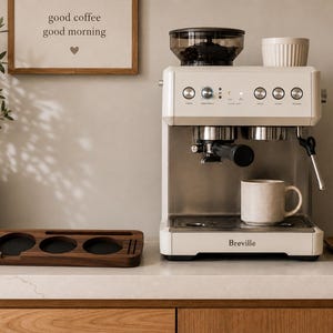 Puede incluir: Una cafetera espresso Breville de color crema con tolva de granos integrada, portafiltro y varilla de vapor. Una taza de cerámica moteada descansa sobre la bandeja de goteo. Junto a la máquina hay un organizador de madera oscura con huecos.