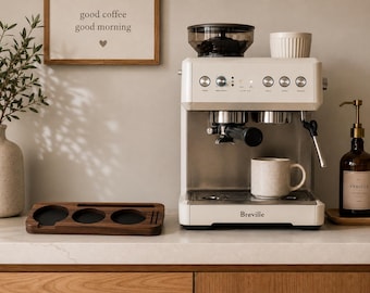 May include: A cream-colored Breville espresso machine with a built-in bean hopper, a portafilter, and a steam wand. A speckled ceramic mug sits on the drip tray. Next to the machine is a rectangular dark wood organizer with three circular cutouts.
