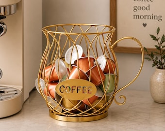 May include: A gold-colored wire coffee pod holder shaped like a large mug with a handle. The front features an oval plaque with the word COFFEE cut out. The holder is filled with various metallic-colored coffee capsules in shades of copper and green.
