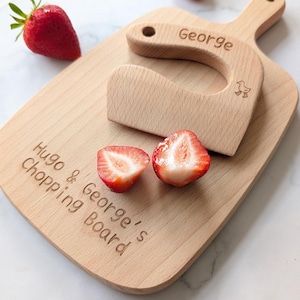 May include: A light-coloured wooden chopping board engraved with the text Hugo & George's Chopping Board, accompanied by a matching wooden cutter engraved with the name George and a small duck icon, alongside two fresh red strawberries.