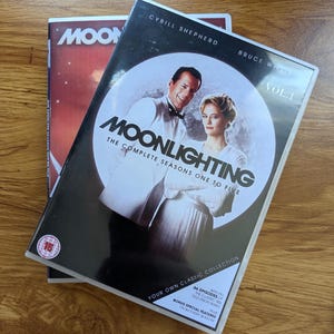 Op de afbeelding: Een Moonlighting The Complete Seasons One to Five dvd-set met Cybill Shepherd en Bruce Willis op de hoes. De zwart-witte hoes bevat tekst over Vol. 1, 66 afleveringen en extra's, met een rood 15-leeftijdsaanduiding symbool.