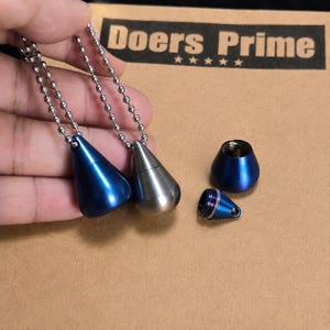 Puede incluir: Dos colgantes de urna de metal en forma de lágrima en cadenas de bolas plateadas, uno azul y otro plateado. Se muestra una parte superior y una base de colgante azul desmontadas. El texto Doers Prime con cinco estrellas está impreso.