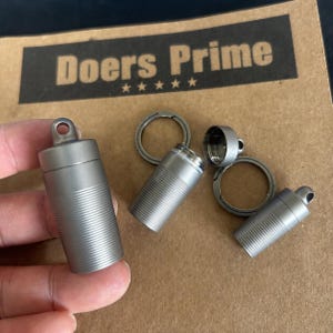 Peut inclure: Trois petits récipients cylindriques gris métallisé avec des couvercles à vis et des boucles intégrées pour la fixation. Chaque récipient présente une fine texture nervurée horizontale sur le corps. Deux sont fermés et un est ouvert.