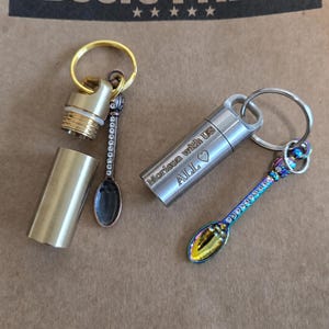 Op de afbeelding: Twee cilindrische metalen sleutelhangerhouders met schroefdeksels en bevestigde miniatuurlepels. De ene is messingkleurig met een donkere lepel, de andere is zilverkleurig met een iriserende lepel gegraveerd met "Morison with Us ALL ♡".