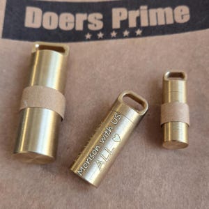 May include: Three cylindrical brass containers with loops at the top. The centre container is engraved with the text "Morison with us ALL" and a heart symbol. Each container features a smooth metallic finish and a small paper band around the body.