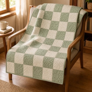 May include: A quilted throw blanket featuring a checkered pattern of alternating sage green and off-white squares. The fabric has a textured, crinkled appearance throughout, and the blanket is draped over a wooden armchair with a light-colored seat.