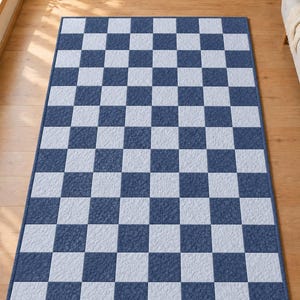 May include: A rectangular area rug featuring a repeating checkered pattern of dark blue and light blue squares. The rug has a finished blue border around the entire perimeter, and the surface of the squares displays a subtle, textured fabric appearance.