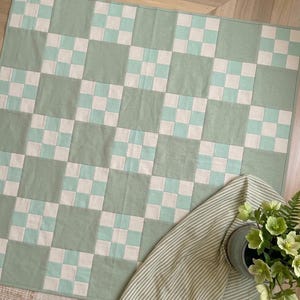 May include: A square quilt featuring a repeating checkerboard pattern of solid sage green squares and smaller nine-patch blocks made of alternating white and sage green squares. The quilt is finished with a thin sage green border around the edges.