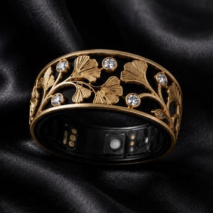 Peut inclure: Une bague connectée dorée présentant un design ajouré complexe de feuilles de ginkgo et de petites pierres claires incrustées. L'intérieur de l'anneau est noir et révèle des capteurs électroniques et des points de contact de charge visibles.