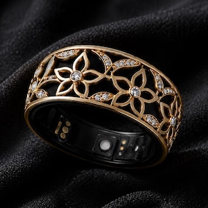 Peut inclure: Une bague connectée dorée présentant un motif floral ajouré complexe sur la bande extérieure. Le design est agrémenté de petites pierres transparentes incrustées. L'intérieur de la bague est noir et montre des capteurs électroniques visibles.