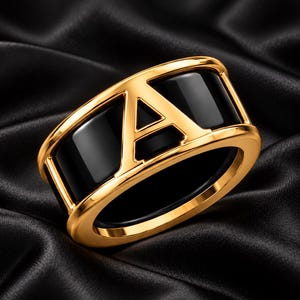 Peut inclure: Une large bague en or ornée d'une lettre majuscule A proéminente au centre. Le design intègre des incrustations en émail noir lisse et brillant qui contrastent avec la finition en métal doré poli, créant une apparence audacieuse et structurée.