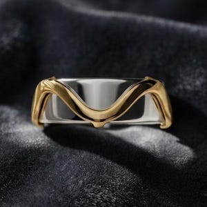 May include: A two-tone ring featuring a polished silver-colored band overlaid with a smooth, undulating gold-colored metal wave design that curves across the center, creating a distinct layered appearance with a high-shine metallic finish.