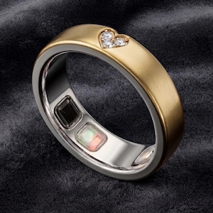 Peut inclure: Une bague connectée avec un extérieur en or brossé présentant une petite découpe en forme de cœur sertie de trois pierres claires. L'anneau intérieur est argenté et contient deux composants électroniques visibles, l'un sombre et l'autre translucide.