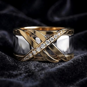 May include: A wide band ring featuring a polished silver-tone center section overlaid with two intersecting gold-tone bands. One of the gold bands is set with a row of small, clear, round-cut stones that sparkle against the metallic finish.