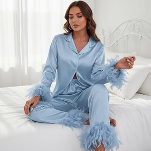 Könnte beinhalten: Ein hellblaues Pyjama-Set bestehend aus einem langärmeligen Hemd mit Knopfleiste und einer passenden Hose. Sowohl die Ärmelbündchen als auch die Hosenbeinsäume sind mit passenden hellblauen Federn besetzt, was für ein einheitliches Aussehen sorgt.