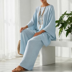 May include: A light blue two-piece set consisting of a loose-fitting, wide-sleeve top and matching wide-leg trousers. The outfit is paired with a long, multi-strand beaded necklace featuring wooden accents and a central circular pendant.