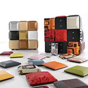 Puede incluir: Dos otomanas cúbicas modulares construidas con paneles cuadrados individuales de diversos colores, texturas y patrones, incluyendo acabados metálicos, sólidos y estampados gráficos. Numerosos paneles cuadrados acolchados están esparcidos en primer plano.