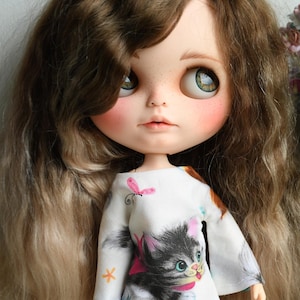 May include: A doll with long brown hair wearing a white shirt with a cartoon kitten and butterflies printed on it.