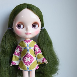 Puede incluir: Una muñeca con el pelo largo verde, que lleva un vestido floral verde y rosa. La muñeca tiene grandes ojos azules con iris morados y rubor rosa en las mejillas.