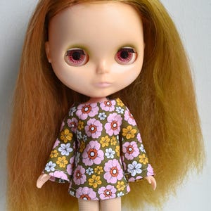May include: A doll with long, light brown hair and large pink eyes. The doll wears a dress with a floral pattern in pink, white, and yellow on a green background. The doll has light skin and is wearing gold boots.