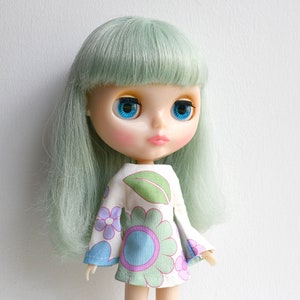 May include: A doll with long, light green hair and blue eyes. She is wearing a white dress with a floral pattern and green leaves.