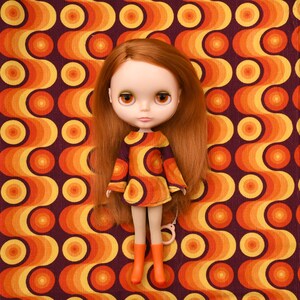 May include: A doll with auburn hair and large brown eyes, wearing a retro-style dress in shades of orange and yellow. The doll is also wearing orange boots. The background features a repeating pattern of orange and yellow circles on a dark red background.