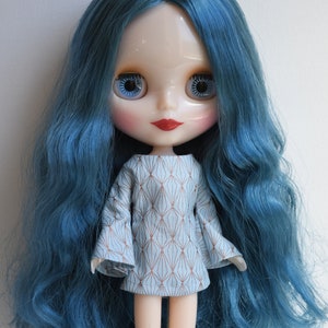May include: A blue-haired doll with blue eyes and red lips wearing a light blue dress with a geometric pattern.