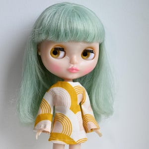 Könnte beinhalten: Eine Puppe mit langen, hellgrünen Haaren, die ein weißes Kleid mit einem gelben und rosa geometrischen Muster trägt.