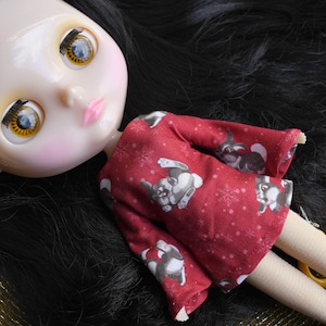 May include: A doll with long black hair wearing a red dress with a white snowflake and grey dog print. The doll has blue eyes with yellow accents.