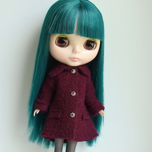 May include: A Blythe doll with teal hair and large eyes is wearing a burgundy coat with three flower-shaped buttons. The coat has a collar and two pockets. The doll is standing in front of a white background.