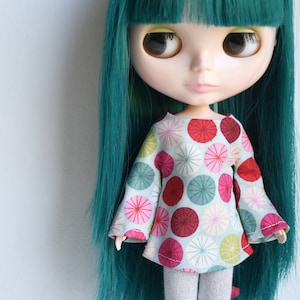 May include: A doll with long teal hair wearing a light blue shirt with a colorful circle pattern. The shirt has long sleeves with bell bottoms.